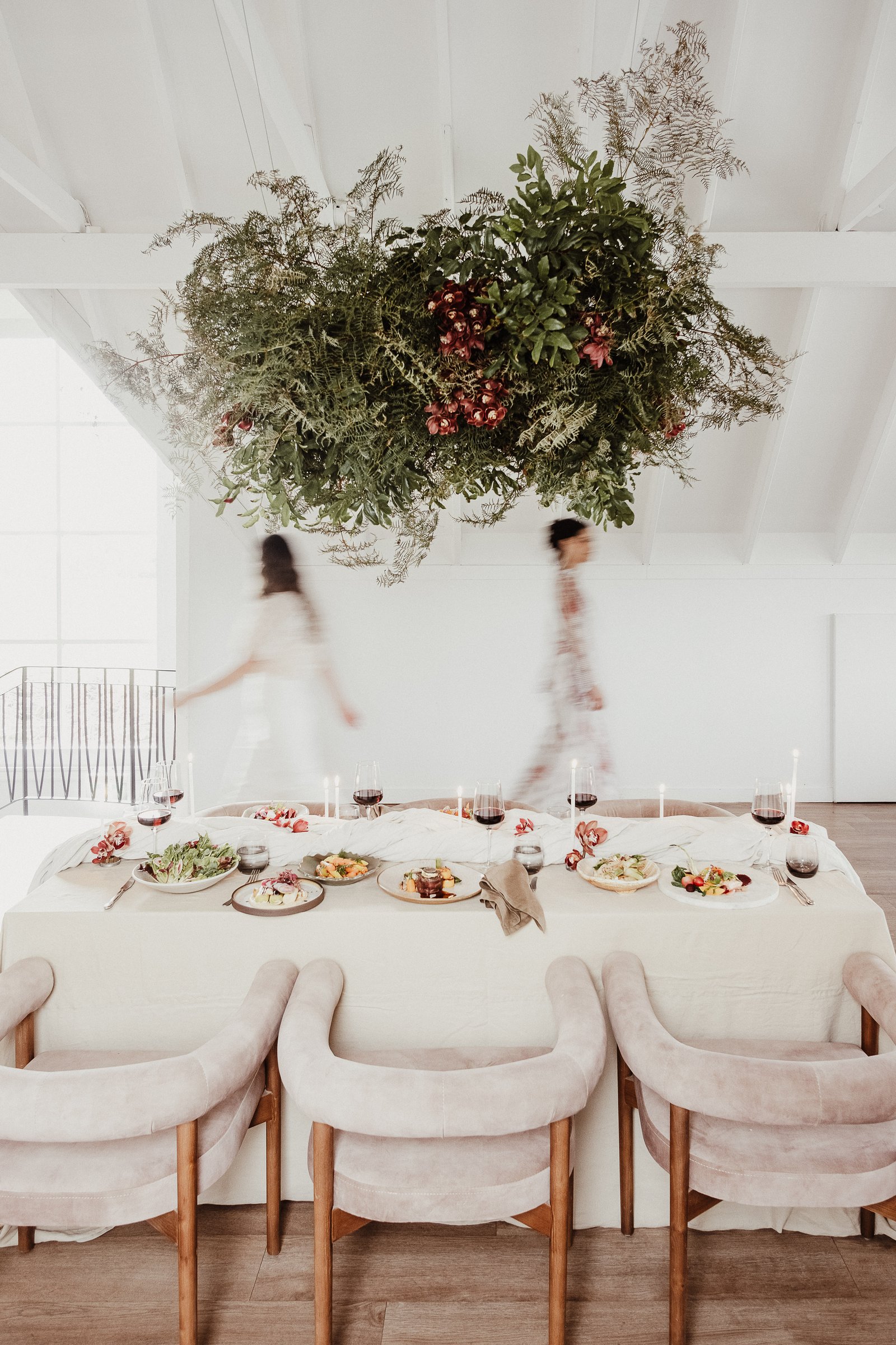 Wedding table beneath a suspended greenery installation, with two brides moving through the bright white venue.