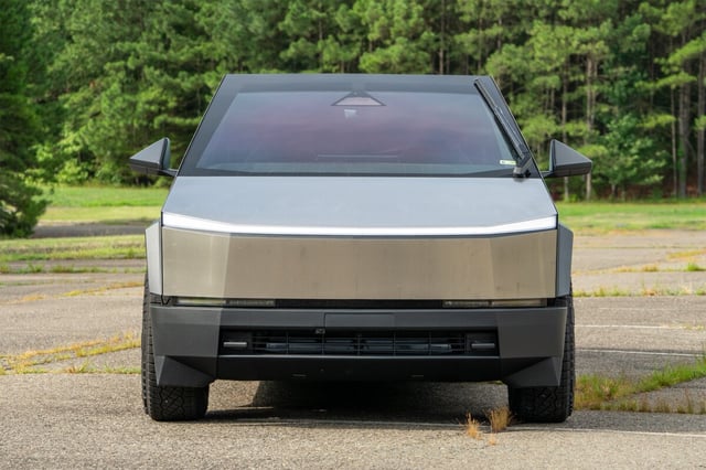 Futuristic, stainless steel electric cybertruck parked on an empty road, surrounded by lush green trees. The front view highlights its distinct angular design and minimalist aesthetic.