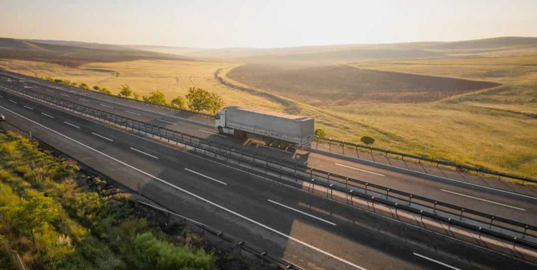 Tarped semi-trailer travelling a divided highway through golden farmland at sunrise, a full truckload en route