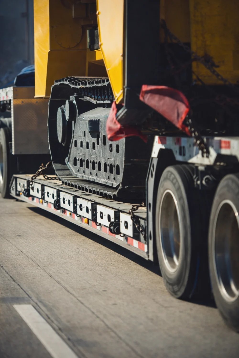 Tracked heavy equipment chained down on a low-deck double drop trailer with red warning flag