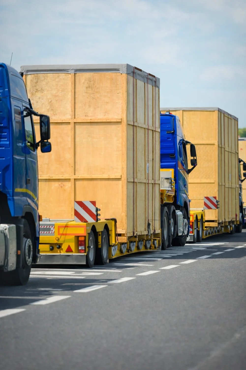 Convoy of semis hauling oversized wooden crates on low-bed double drop trailers with hazard striping
