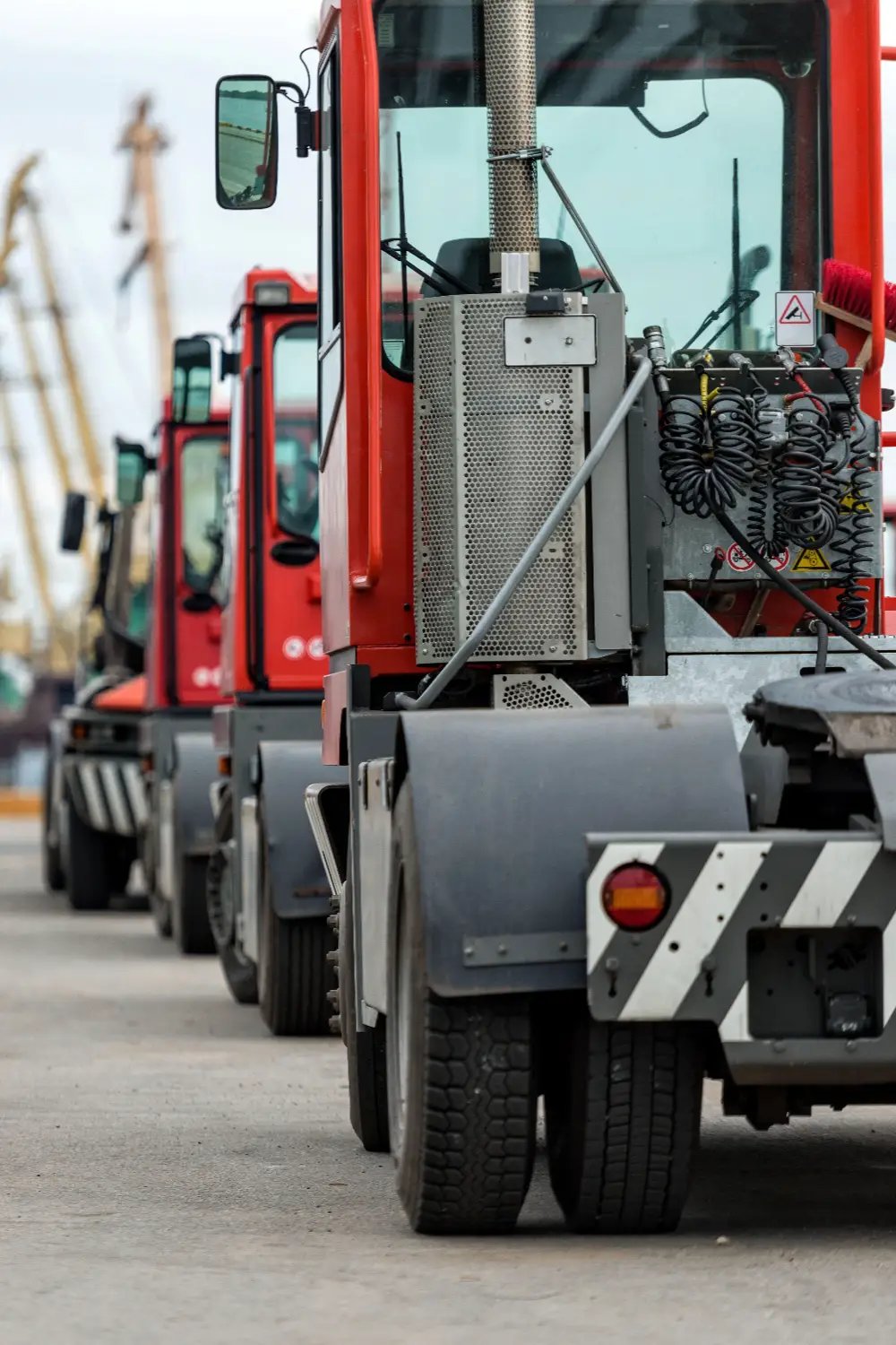 Red terminal tractors lined up at a port container yard with cranes in the background
