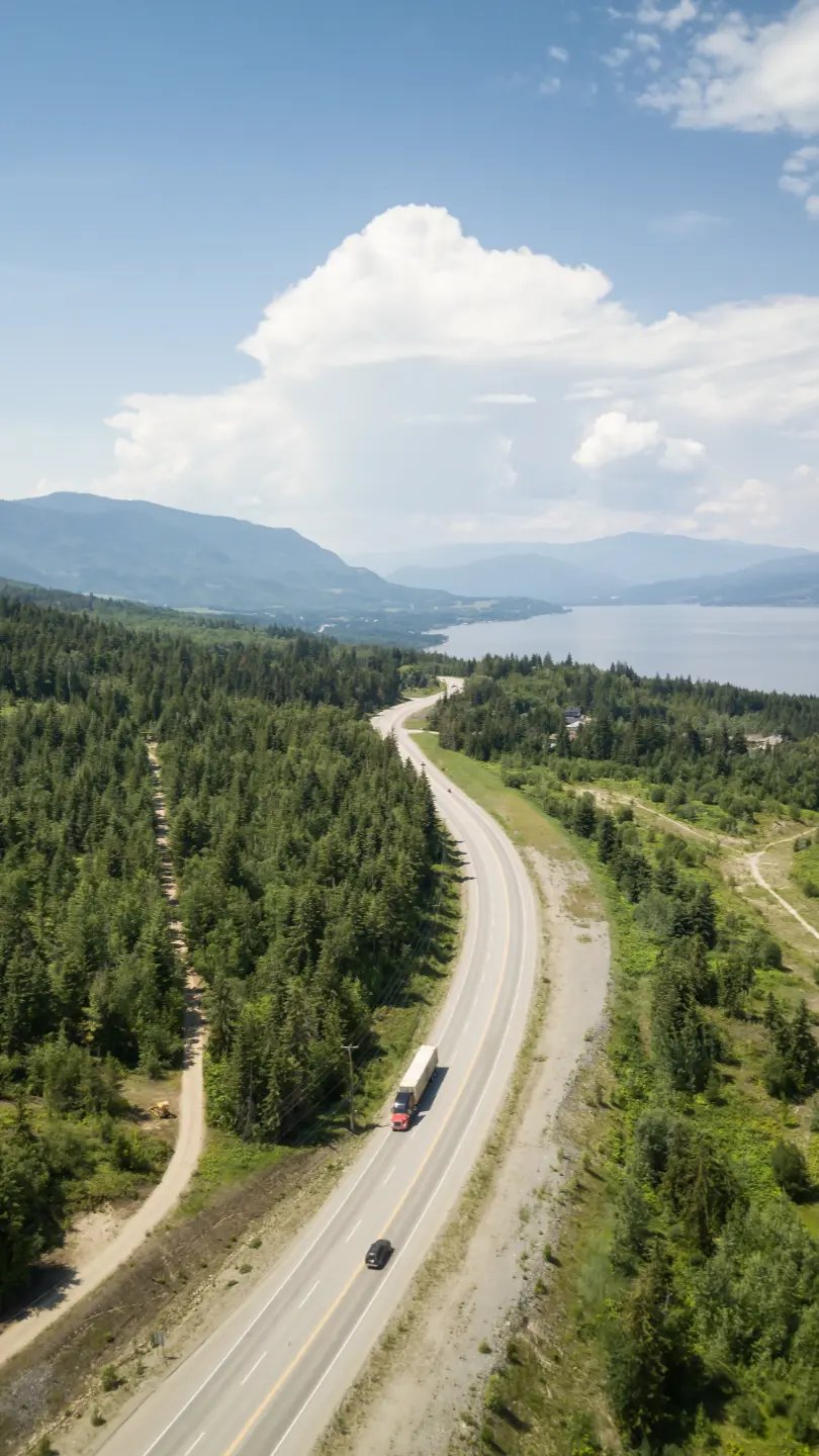 Aerial view of a semi truck on the Trans-Canada Highway winding past a lake and forested mountains in BC