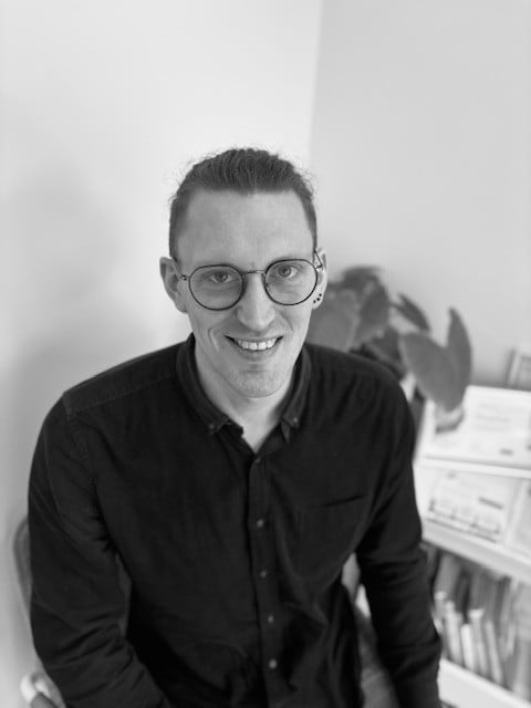 Nick Headshot - The Audiology Place portrait of a smiling man with round glasses wearing a black button down shirt in an office setting nick headshot audiologists dr signe steers charlen lavoie dr nick morrison