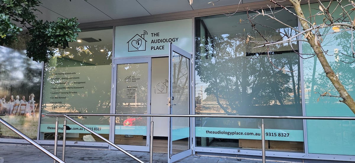 front of the audiology place clinic with glass doors logo of a house with an ear and tree reflections in the windows railing in foreground 2026 08 01 audiologist epping the audiology place