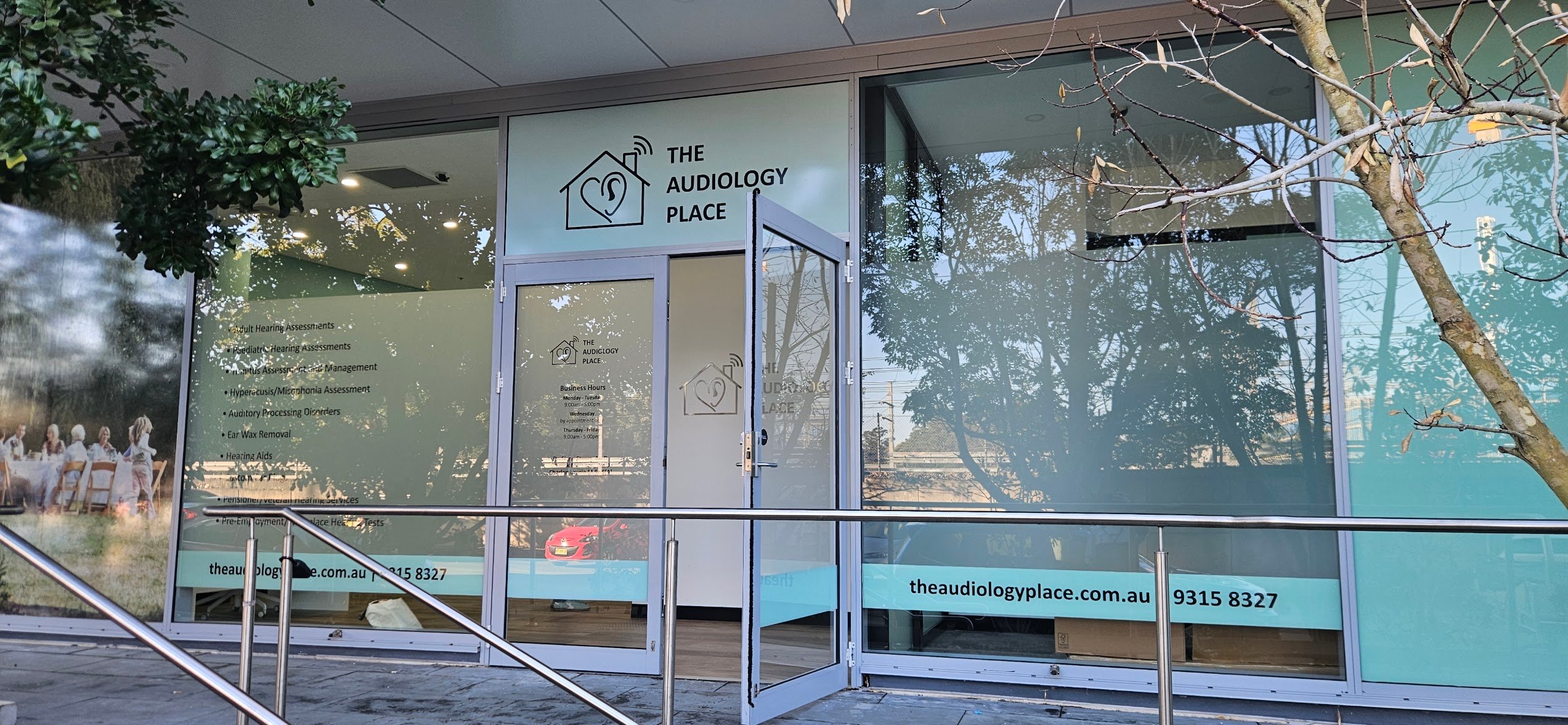 front of the audiology place clinic with glass doors logo of a house with an ear and tree reflections in the windows railing in foreground 2026 08 01 independent audiologist epping the audiology place