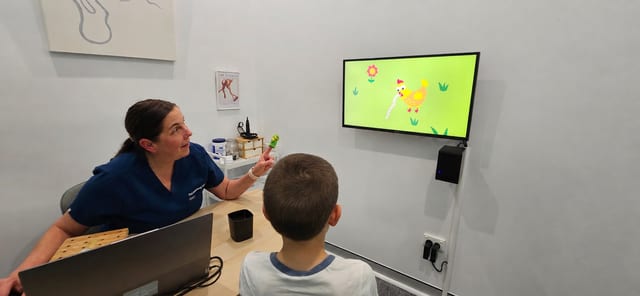 VROA - The Audiology Place healthcare professional in blue scrubs sits at a table with a laptop pointing at a green finger puppet while a child watches a cartoon chicken on a wall monitor vroa visual reinforcement orientation audiometry vroa how we test hearing in babies and toddlers the audiology place