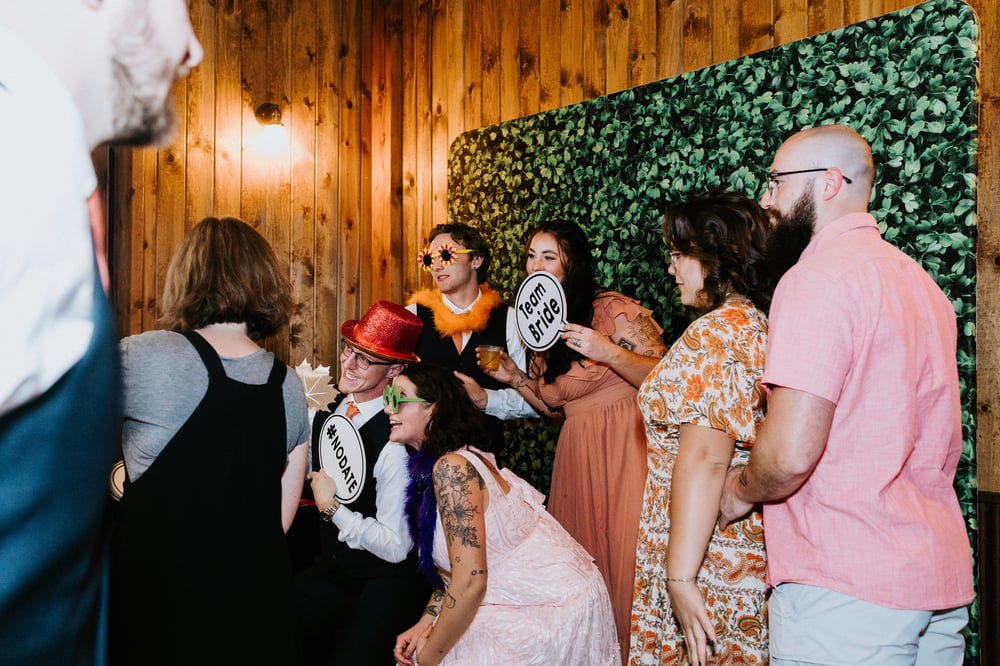 Guests with props at Firefly's open-air photo booth, greenery backdrop, at an Orchard River View wedding — Photo: Kimberly Crist