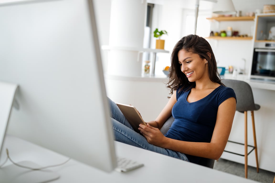 Fotografía de una mujer joven de tes latina trabajando en casa que viste una playera color azul marino y se encuentra sentada frente al monitor de su computadora.