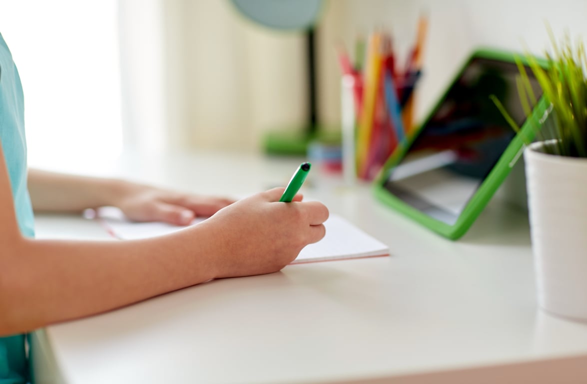En la fotografía aparecen los brazos de una niña en clases en línea que escribe sobre el pupitre de su casa con una tableta electrónica al frente.