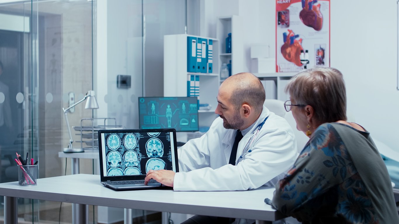 Fotografía en la que aparece un doctor mostrando una tomografía a una señora de edad adulta dentro de un consultorio color blanco.