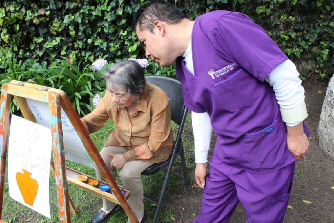 Fotografía en donde se aprecia a una señora de la tercera edad dibujando frente a un caballete mientras es orientada por un cuidador de mediana edad que lleva puesto uniforme de enfermero color morado. Créditos a Beneficiarios de Alzheimer México