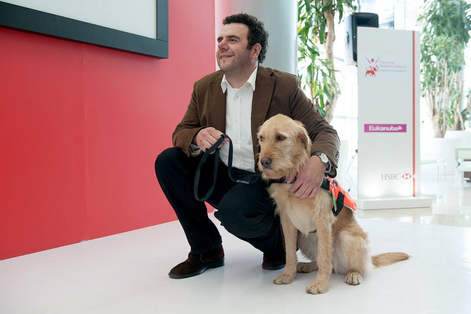 Fotografía de Juan Serrat, un hombre de mediana edad que utiliza un saco café, camisa blanca y pantalón negro, se encuentra hincado a lado de Leire, un perro perro de asistencia de raza labrador de color beige
