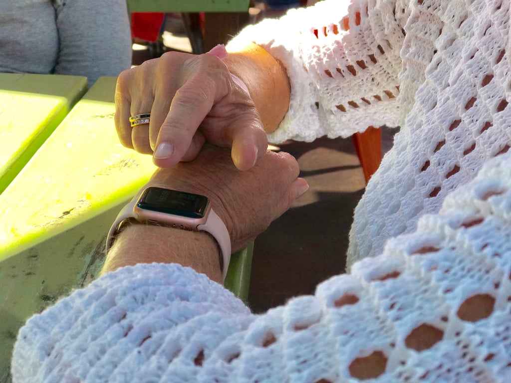 Fotografía de las manos de una señora sobre una mesa de madera color verde, la señora lleva puesta una blusa blanca y tiene su dedo sobre un apple watch, un reloj inteligente con correa rosa.