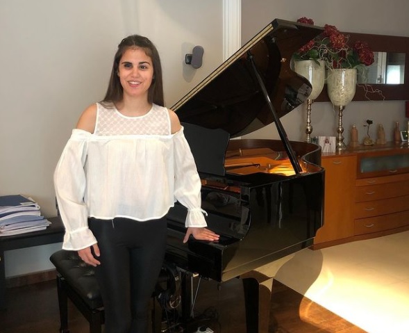 Fotografía de Laura Diepstraten, una mujer joven que aparece frente a un piano de cola color negro, ella va vestida con una blusa blanca con manga ancha y hombros descubiertos y un pantalón negro, tiene el cabello color castaño, ojos cafés, sonría frente a la cámara.