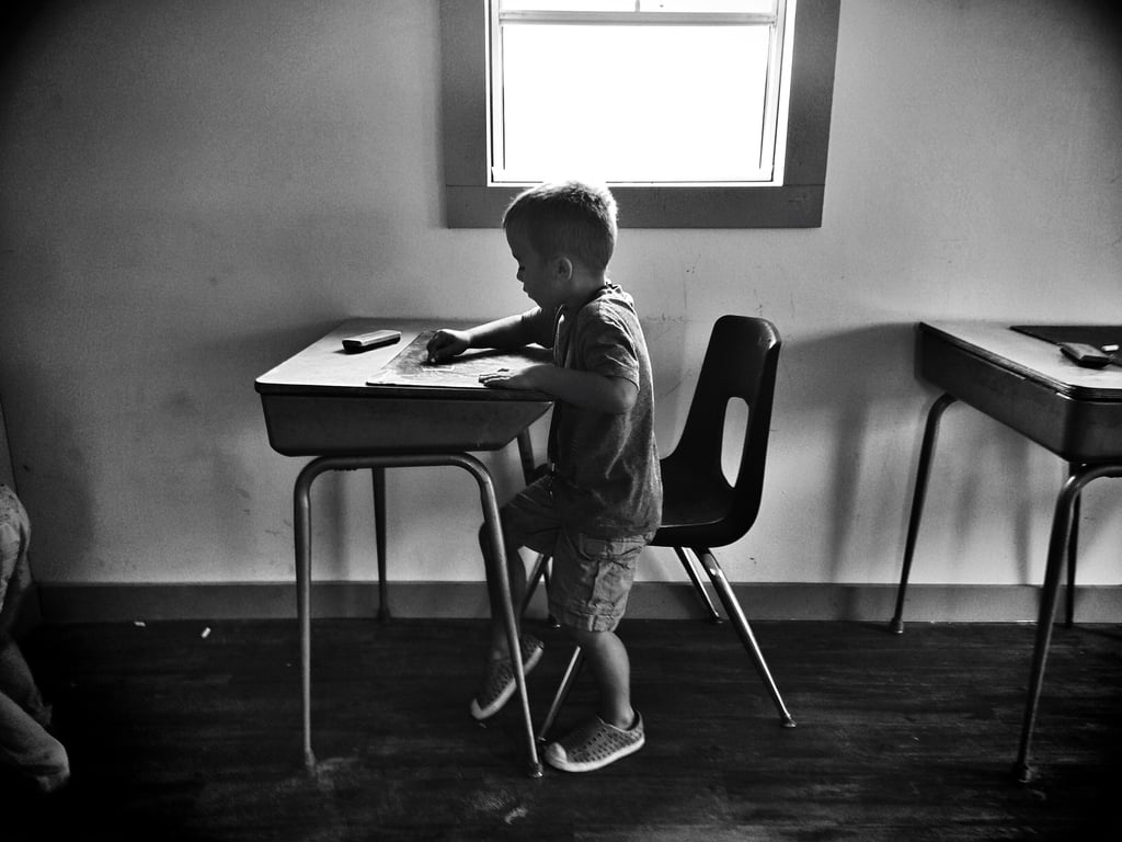 Fotografía a blanco y negro de un niño sentado en una banca escolar, coloreando sobre su escritorio, se encuentra frente a ventana.