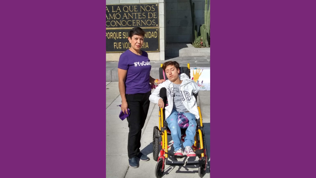 Fotografía de una mujer adulta de pie, vestida de pantalón negro y blusa morada a lado de una joven en silla de ruedas con pantalón de mezclilla azul y suéter blanco.