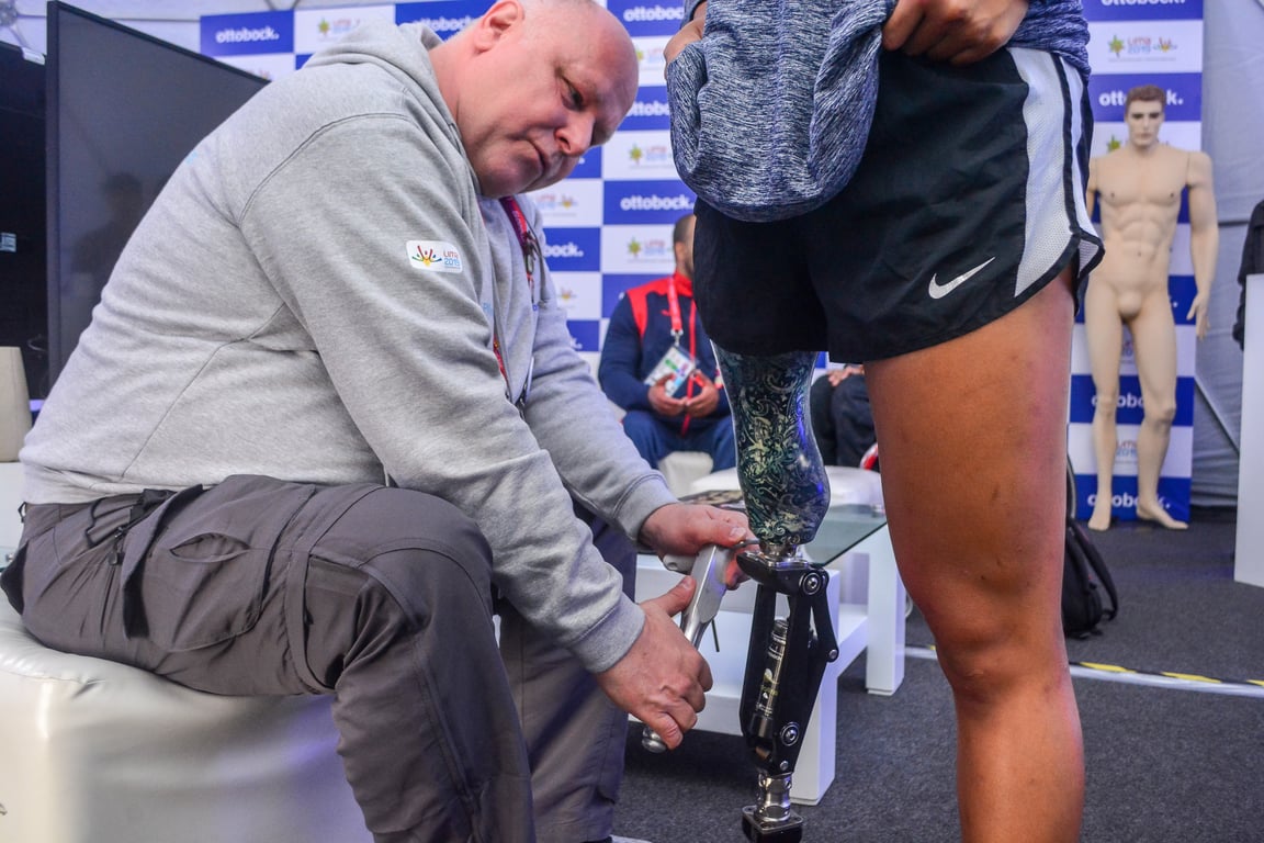 Fotografía de Thomas Pfleghar, un señor maduro con cabello cano corto que viste una sudadera de color gris y bermudas de color gris oscuro, de pie frente a él, se alcanza a apreciar la pierna izquierda de un deportista con short negro de la marca Nike y una prótesis en la pierna derecha.