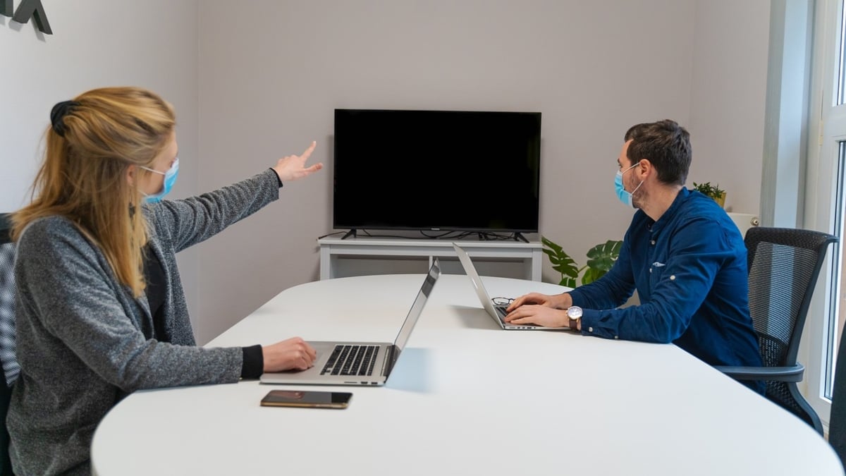 Fotografía de Pixabay en la que se aprecia a dos personas: una mujer, a la izquierda, y el hombre, a la derecha, trabajando en una mesa grande, color blanco, en una sala de oficina. Para seguir medidas sanitarias y evitar contagios, ambos usan cubrebocas, que se aprecian aunque en la imagen están de perfil. Además, no están sentados frente a frente, sino que dejan un espacio entre ellos. Los dos tienen laptops abiertas, son de color plateado. La mujer, también, tiene un celular sobre la mesa, al lado de su computadora. Ambos voltean hacia una pantalla que está al fondo de la sala donde están y la mujer la señala con la mano izquierda. Los dos visten ropas en color que van del gris al azul marino. Ella tiene cabello largo, rubio y semi recogido, y él tiene cabello corto, oscuro.