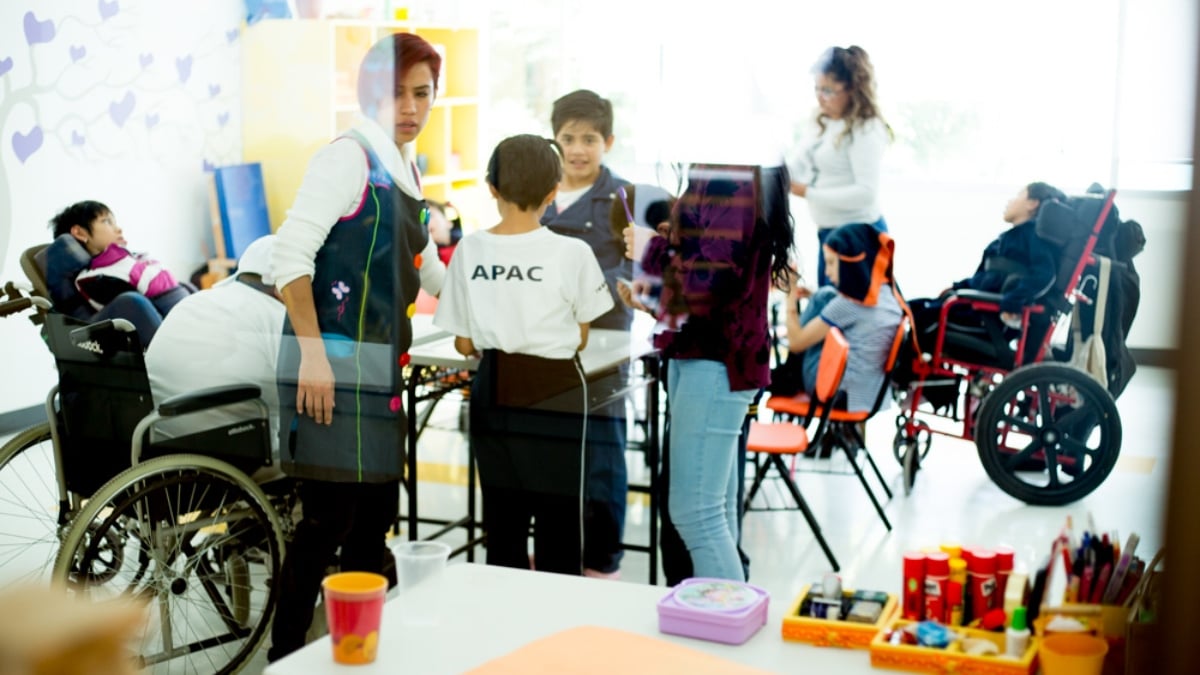 Fotografía de uno de los salones de clases de APAC, donde se ven algunas de las terapeutas y maestras con un grupo de niños y niñas con parálisis cerebral. La fotografía es a distancia y no se ven los detalles de cada persona, aunque sí se aprecia a uno de los niños que está de espaldas, al centro, y viste una playera blanca que dice APAC. Algunos de los niños están en sus sillas de ruedas y el único que ve a la cámara sonríe abiertamente.