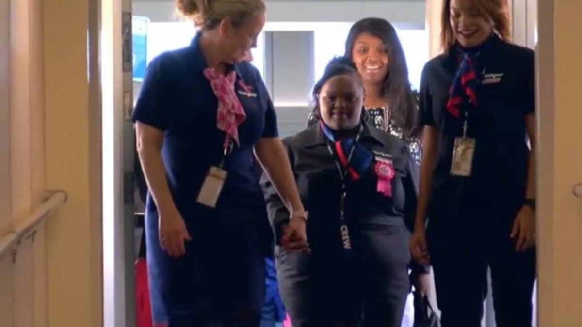 Fotografía de un grupo de asistentes de vuelo de American Airlines, que visten su tradicional uniforme azul oscuro con una pañoleta rosa al cuello. En el centro está una joven afroamericana que tiene síndrome de Down, y que recibió un contrato honorario de la aerolínea para que apoye a su personal en cada vuelo que realice.