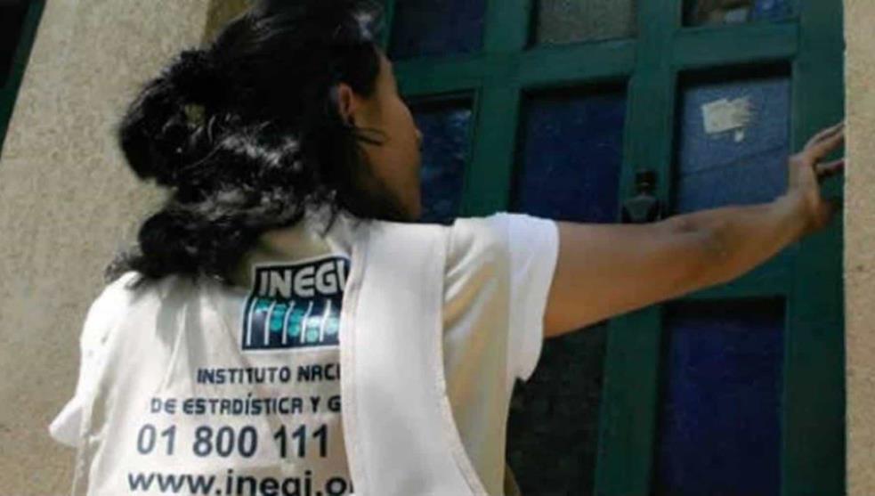 Fotografía de una mujer de cabello color negro que se encuentra de espaldas a la cámara, aparece tocando el tiembre y frente a ella una puerta de metal de color verde olvivo, la mujer lleva puesto un chaleco de color café claro que dice en letras azules la palabra “INEGI”.