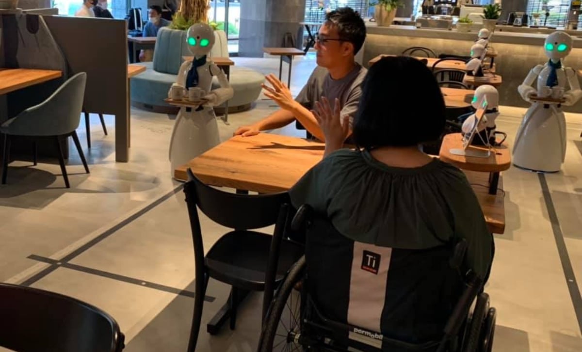 Fotografía de dos personas, una mujer de espaldas usuaria de silla de ruedas, y un hombre sentado de tres cuartos, frente a una mesa de un café en la ciudad de Tokio, que es atendido por robots humanizados operados a distancia por personas con discapacidad. Al lado del hombre se ve uno de los robots, vestido de blanco, con una corbata negra y grandes ojos que se iluminan con una luz verde. El robot lleva una charola. Otro robot está del otro lado de la mesa de las dos personas sentadas a la mesa.