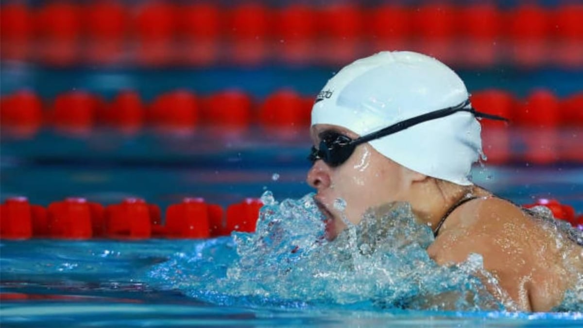 Fotografía de Naomi Somellera, la campeona paralímpica mexicana de natación. En la imagen, se le ve de perfil, nadando en una alberca con carriles rojos. Naomí lleva gorro de natación blanco y sobre él sus goggles negros. La fotografía fue captada justo en el momento en el que jala aire y en torno a ella se ve el agua de la alberca agitada, como en una pequeña ola.