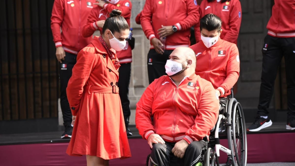 Fotografía de Ana Guevara y el campeón parapanamericano de Powerlifting José de Jesús Castillo, conversando antes de la ceremonia de abanderamiento de la delegación de México que estará en Tokyo 2020. La imagen muestra a una mujer que conversa con un hombre, sin cabello, usuario de silla de ruedas. Atrás de ellos se alcanzan a ver otros paradeportistas. Todos visten el uniforme de México que consta de chamarra color rojo, con la bandera nacional en el lado izquierdo del pecho, y pants negros. Los dos llevan cubrebocas blanco, también con la bandera nacional a un costado.