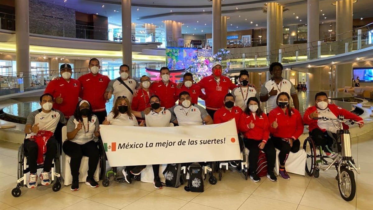 Parte de la delegación mexicana que asiste a los Juegos Paralímpicos de Tokyo 2020. Son 18 personas colocados en dos filas, todos con el uniforme del equipo que consta de pants negros y chamarra roja. Todos usan cubrebocas y despliegan un letrero que dice "México la mejor de las suertes", entre las banderas de México y Japón.