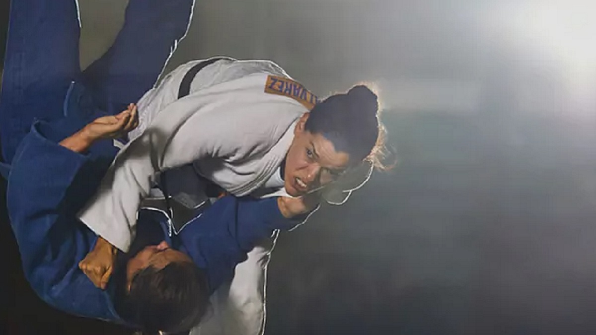 Imagen de dos judocas mujeres que tienen baja visión. Una de ellas viste un traje en color azul y está suspendida en el aire, con las piernas y la cabeza hacia arriba; la otra deportista tiene un traje blanco y sostiene a su oponente en el aire, con una llave.