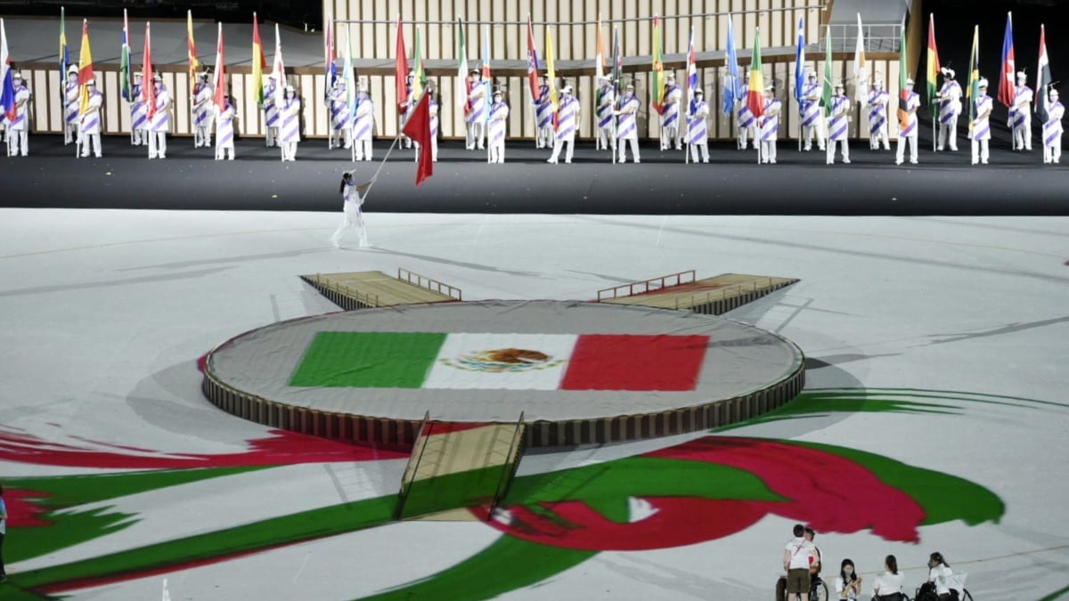 Fotografía de un momento de la inauguración de los Juegos Paralímpicos Tokyo 2020. En la parte posterior están dos filas de personas con banderas de los países participantes y al centro del escenario se ve la bandera mexicana, rodeada de gráficos en colores verde, blanco y rojo.
