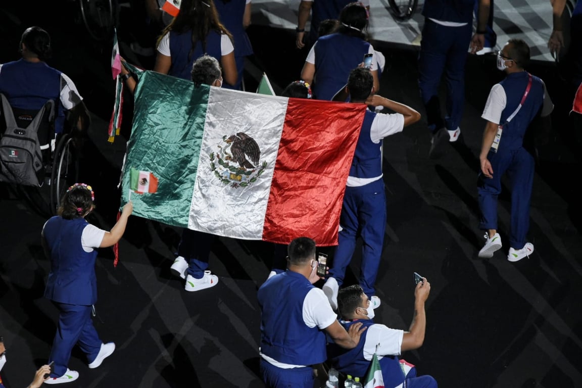 Fotografía que muestra una enorme bandera de México entre miembros de la delegación que representa al país en los Juegos Paralímpicos de Tokyo 2020. Los y las atletas que desfilan visten el uniforme azul con playera banca, y fueron captados de espaldas.