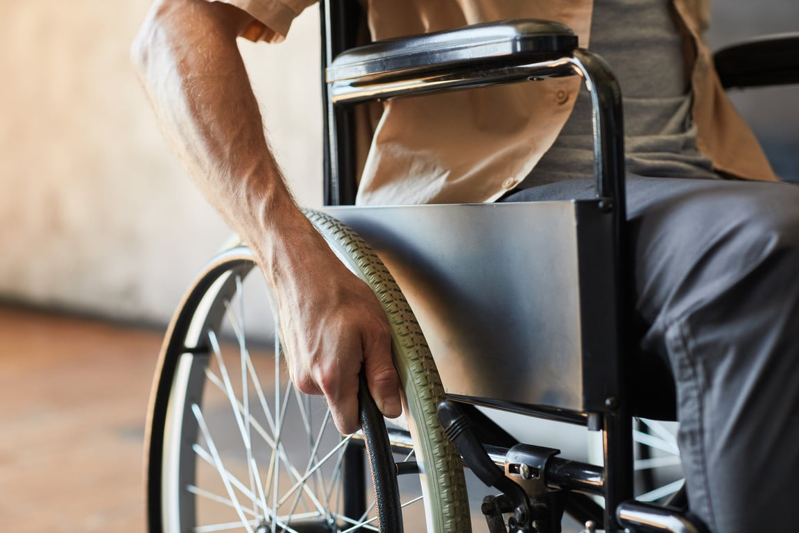 Fotografía en donde se alcanza a apreciar brazo izquierdo de una persona usuaria de silla de ruedas que toca la rueda de la silla color negra, la persona lleva puesta una camisa color beige con playera gris y pantalón negro.