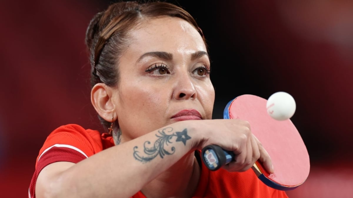 Fotografía de Edith Sigala, deportista de tenis de mesa durante su participación en Tokyo 2020. En la imagen se ve a una mujer madura, de cabello café, peinado en un chongo impecablemente elaborado que combina trenzas y cortes que terminan en la parte alta de la cabeza. Tiene ojos enormes, maquillados y las cejas delineadas. La posición de su cara es de tres cuartos y casi a la altura de su boca cruza su brazo derecho, en el que destaca un tatuaje de estrellas en la muñeca, y su mano sostiene una raqueta o paleta de tenis de mesa en color rojo, con mango negro, mientras golpea una pelota blanca. La actitud de la jugadora es de concentración. La imagen, casi un close-up, permite ver que la deportista viste la camiseta roja del uniforme de la delegación mexicana.