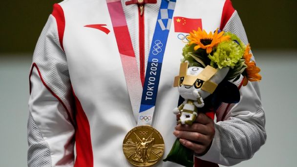 Fotografía del pecho de una persona vestida con una chamarra blanca con franjas rojas a los costados que tiene impresa la bandera de china, la persona sostiene un ramo de flores con la mano izquierda y lleva colgada una medalla de oro del cuello, el listón de la medalla tiene impreso “Tokio 2020”.