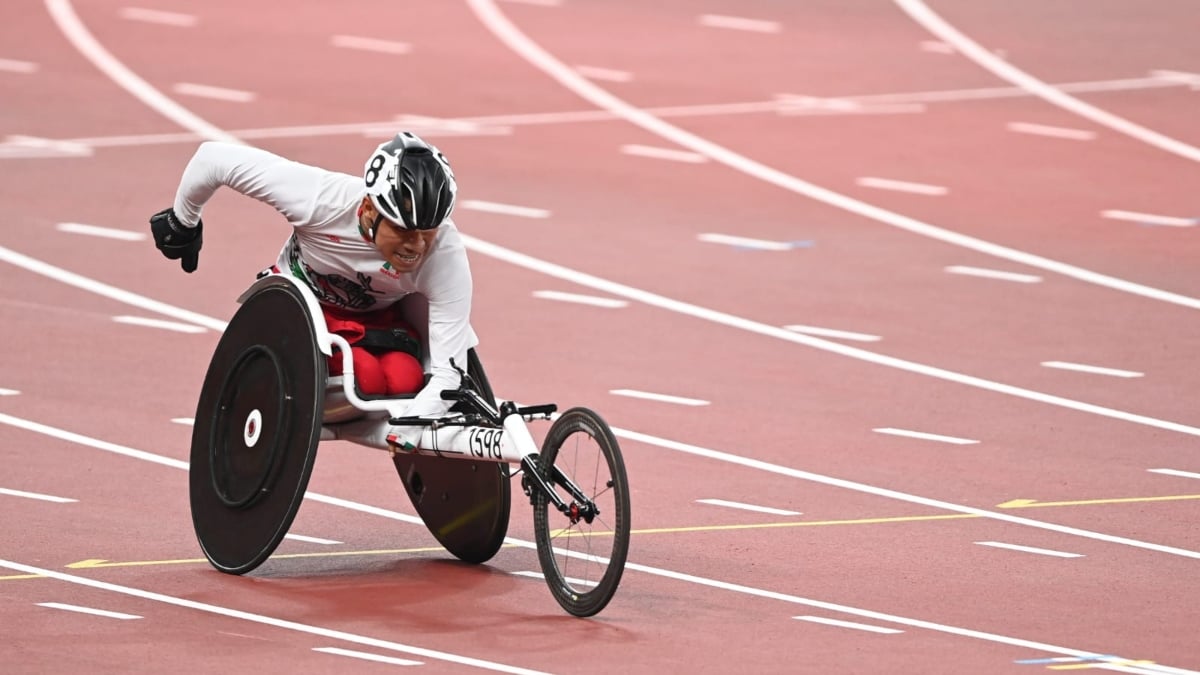 Fotografía del atleta mexicano Leonardo Pérez, quien es usuario de silla de ruedas y compite en carreras de velocidad con un equipo adaptado. En la imagen se le ven en la pista de tartán color rojo con carriles marcados en blanco, saliendo de una curva. El ángulo de la imagen permite ver al competidor, que viste playera de competencia blanca con un casco de protección a juego, con la cabeza inclinada y el brazo derecho en alto, para impulsar su movimiento, mientras con el brazo izquierdo controla la silla de carreras.