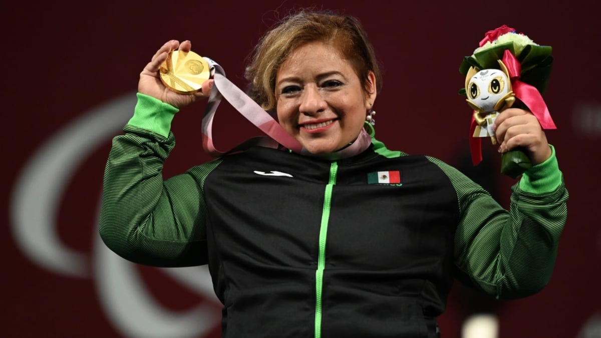 Fotografía de la campeona mexicana Amalia Pérez. La mujer, de mediana edad, posa sonriente mientras muestra la medalla de oro que ganó en la competencia de powerlifting en Tokio 2020. Ella está sentada en su silla de ruedas, viste pants negros con las mangas y el cierre delanteros color verde, con la bandera mexicana en el lado superior derecho. Tiene el cabello corto, teñido de color café rubio, más corto del lado izquierdo y peinado de lado. En el brazo izquierdo muestra el arreglo floral que entrega a los ganadores de medallas, y con la derecha jala el cordón que sostiene la medalla dorada que cuelga de su pecho y la alza a la vista de todos.