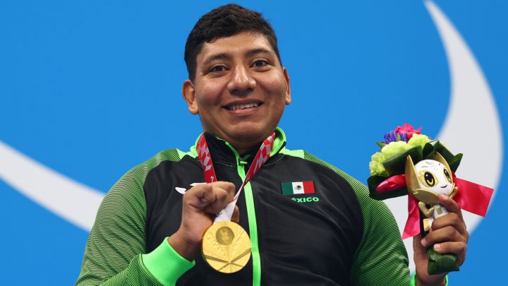 Fotografía de Jesús Hernández, nadador paralímpico que ganó la medalla de oro. En la imagen está sentado en su silla de ruedas, en el podio donde le entregaron la presea. Su silla de ruedas es negra, mismo color de los pants que usa y en el que destaca el color verde de las mangas de la chamarra. Es un hombre de 29 años, de cabello corto y negro, que usa un cubrebocas blanco. En su pecho luce la medalla de oro, y con la mano izquierda sostiene el arreglo de flores que le fue entregado como medallista. Atrás de él, en una pared azul está el emblema de los Juegos Paralímpicos de Tokyo 2020 en color blanco.