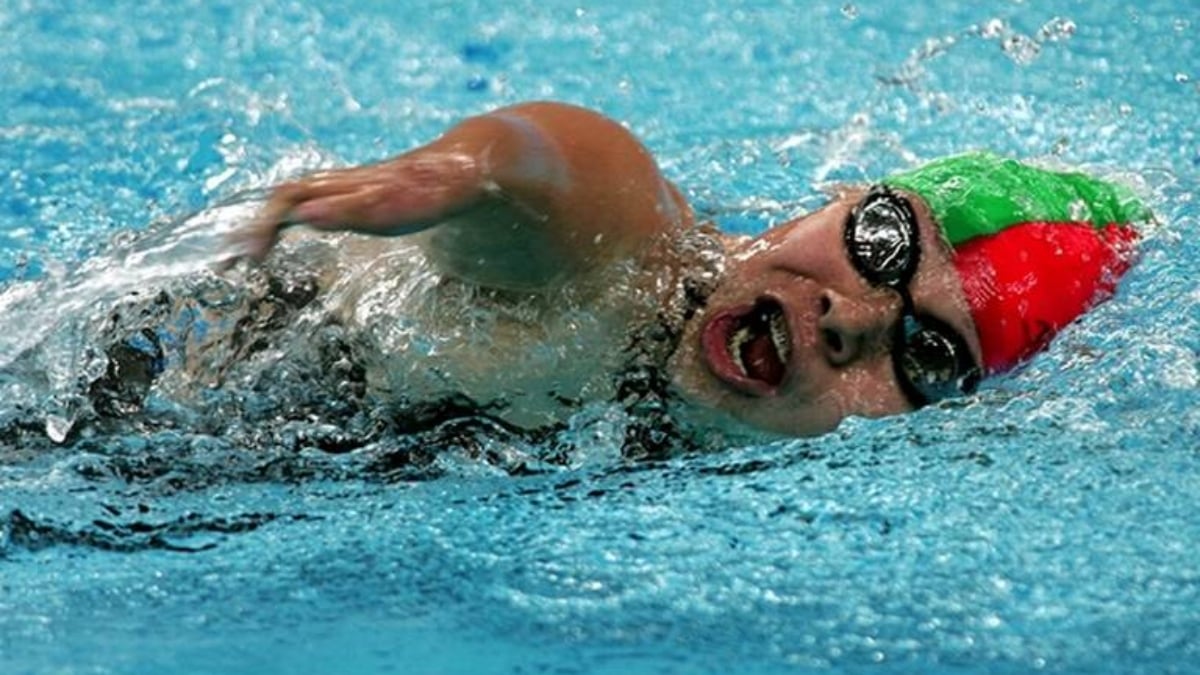 Fotografía de la sirena mexicana Patricia Valle, quien impuso un récord al participar en siete Juegos Paralímpicos. La imagen capta a la nadadora dentro de la alberca, con la cara de lado para respirar, tiene la boca abierta, goggles negros, y gorro de natación en dos colores: verde y rojo.