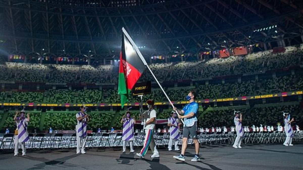 Momento en que un hombre desfila con la bandera de Afganistán en la ceremonia de apertura de los Juegos Paralímpicos de Tokyo 2020, aunque la delegación de dos atletas canceló su participación por la situación política en ese país. En la imagen, el abanderado vestido de gris y azul sostiene la bandera verde con rojo, entre una valla de voluntarios vestidos de blanco, que no se aprecian bien debido a la distancia de la foto.