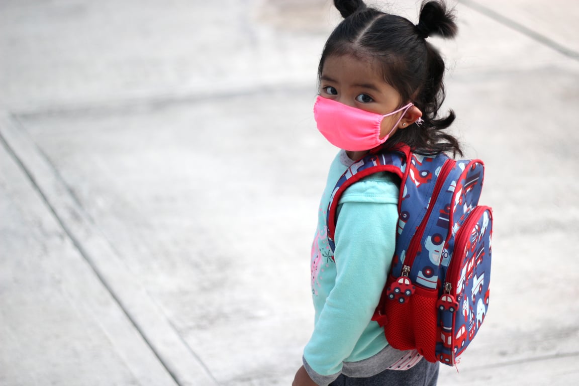 Fotografía de una niña de tez morena, pequeña, de cabello rizado quebrado de color castaño oscuro, peinado de dos coletas, sudadera color azul, con cubrebocas de color azul claro, tiene colgada en sus hombros una mochila azul con bordes de color rojo, se encuentra de pie hacia al frente, pero girando ligeramente para ver directo a la cámara.