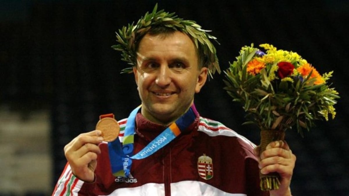 Fotografía del atleta húngaro Pál Szekers, único que ha ganado medalla olímpica y paralímpica. En la imagen se ve a un hombre de nariz recta y larga, rubio, que sonríe tras recibir una de las medallas que ganó, y a la que muestra levantándola con la mano derecha. En la cabeza tiene una corona de laurel en señal de victoria, y en la izquierda un ramo de flores.