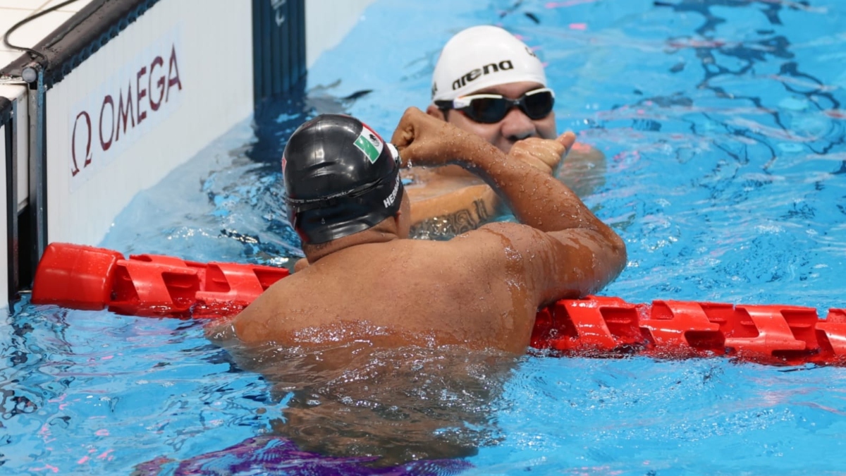 Fotografía de los campeones paralímpicos Arnulfo Castorena y Jesús Hernández, tras un histórico triunfo en Tokyo 2020. Ambos celebran dentro de la alberca. Hernández da la espalda y se ve como abraza a Castorena, quien ganó el oro al llegar en primer lugar. Hernández usa una gorra plástica color negro, con la imagen de la bandera mexicana en el lado derecho. Castorena usa una gorra blanca, en la que se aprecia parte de su apellido y aún trae puestos unos goggles negros.