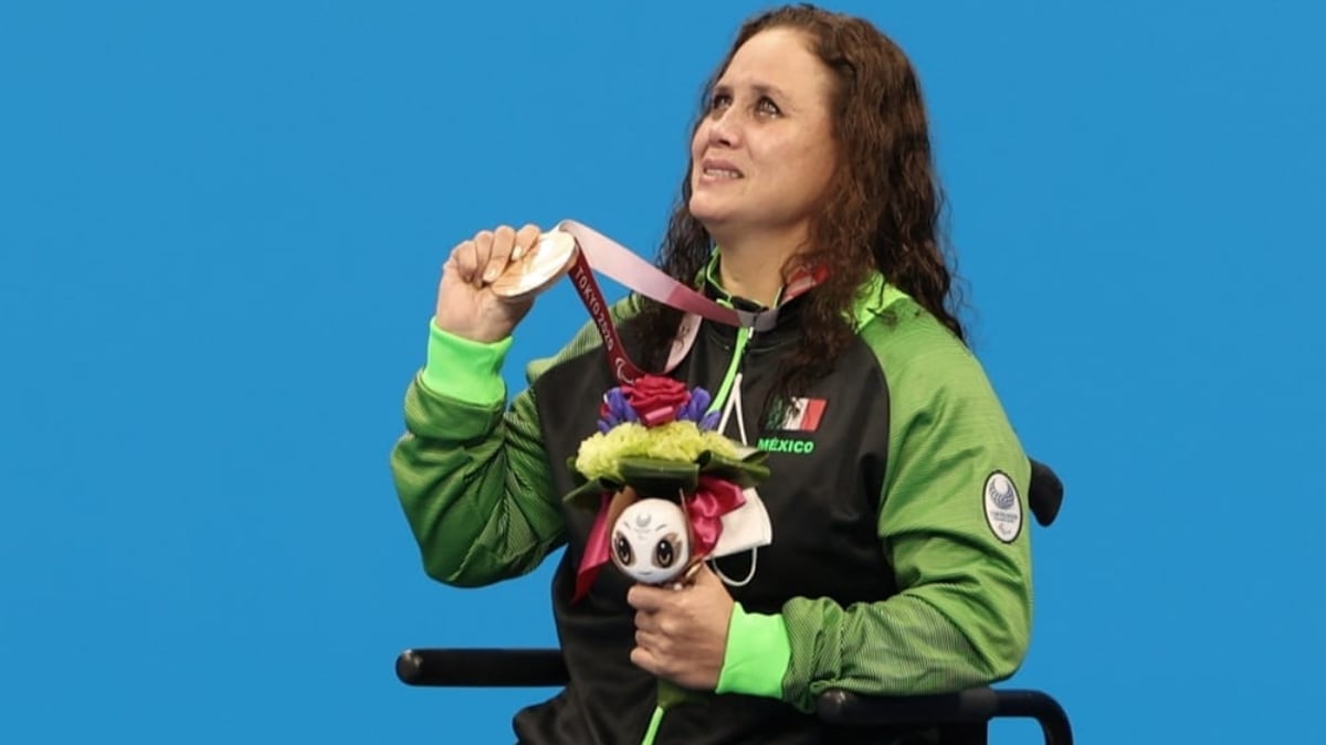 Fotografía de Nely Miranda, nadadora paralímpica. Ella es una mujer madura, usuaria de silla de ruedas. La fotografía tiene un encuadre de tres cuartos, que permite apreciarla cuando levanta y muestra la medalla que cuelga en su cuello con la mano derecha, mientras la izquierda permanece pegada a su cuerpo sosteniendo un ramo de flores. Ella tiene el cabello largo, semirrizado, teñido de color caoba, y viste el uniforme de la delegación mexicana, aunque solo es visible su chamarra negra con mangas verdes.