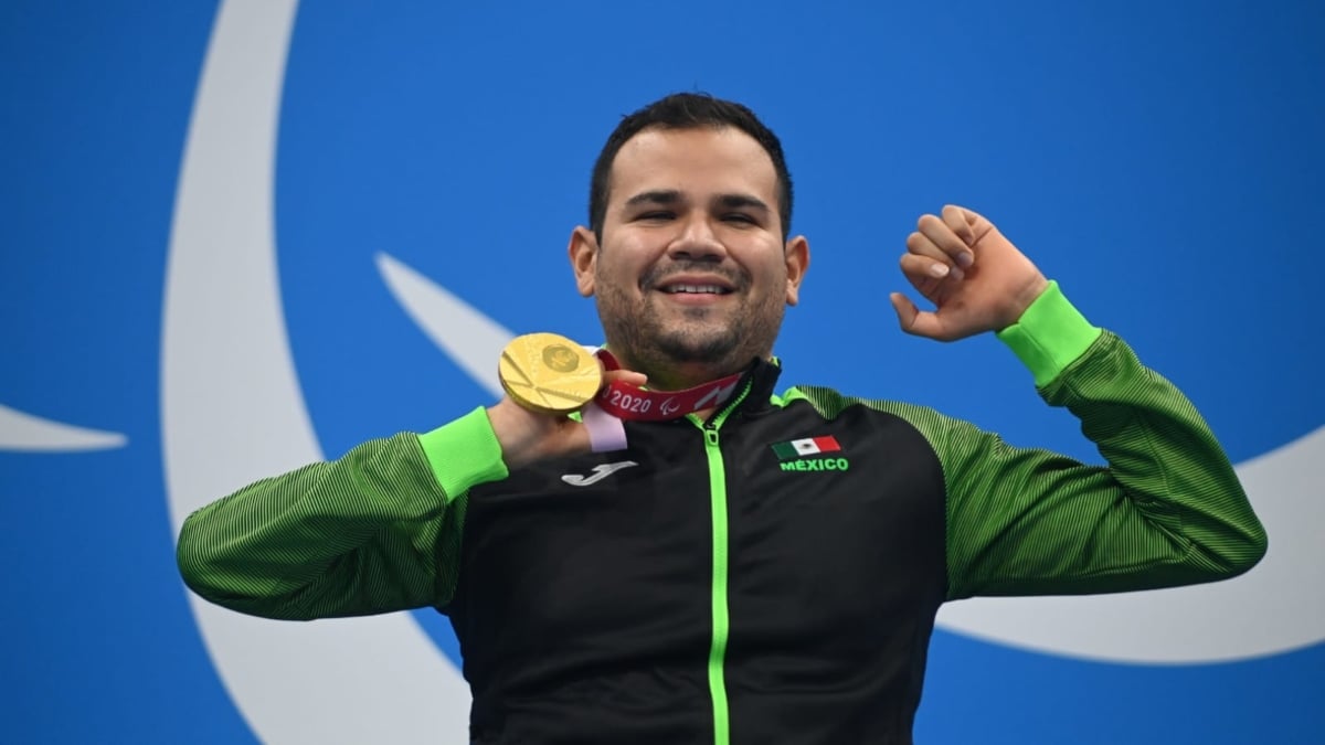 Fotografía de Diego López, medallista de oro en Tokio 2020. Él es usuario de sillas de ruedas, aunque en la imagen solo se ve la mitad superior de su cuerpo. Es un hombre joven, de cabello oscuro, muy corto, que sonríe ampliamente mientras muestra su medalla de oro en natación paralímpica con la mano derecha, la otra mano la empuña y sube a la altura de su cabeza. Viste la chamarra oficial del equipo mexicano que es negra con mangas y el cierre verde.