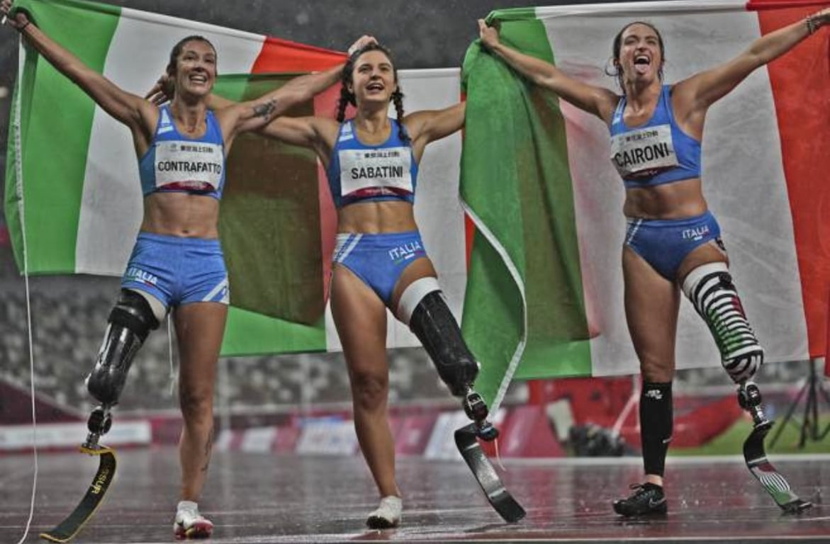 Fotografía de las tres atletas italianas que hicieron el 1, 2, 3 en la carrera de 100 metros. Las tres son mujeres jóvenes, que tienen la amputación de una pierna -dos de ellas la izquierda y la otra la derecha- y llevan prótesis hasta el muslo. Las tres visten uniforme de competencia, color azul claro, el top es muy corto y el short es corto. Cada una de las jóvenes tiene una bandera de Italia, verde, blanca y roja, y la pasan por atrás de su cabeza mientras alzan los dos brazos.