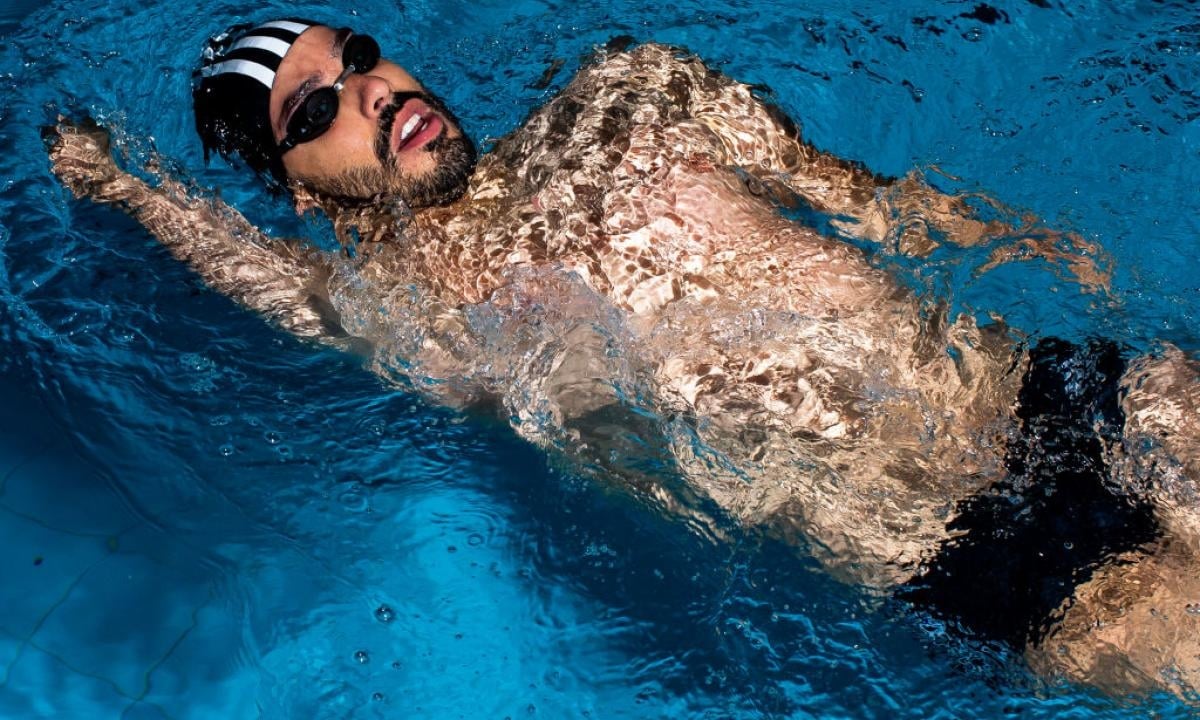 Fotografía de Daniel nadando de dorso. Lleva barba y bigote negros, goggles negros y un gorro negro con francas verticales blancas.