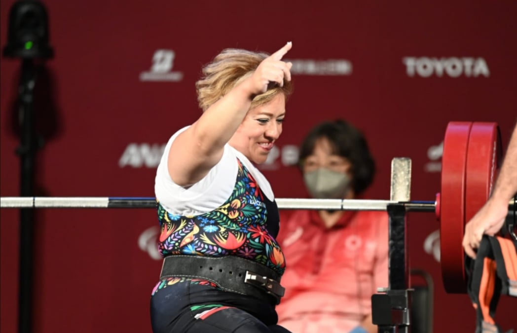 Fotografía de Amalia Pérez, una mujer de mediana edad, robusta, de tez morena clara, cabello teñido de rubio, está de perfil frente unas pesas con barras plateada y discos color rojos. Sonríe mucho y levanta su brazo y dedo índice.