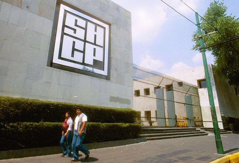 Fotografía del edificio de la Secretaría de Hacienda y Crédito Público, una pared de color gris con letras de gran tamaño con “SHCP” hechas de metal, sobre la acera de enfrente caminan dos personas, una mujer y un hombre, la mujer viste pantalón de mezclilla de color azul, blusa de manga larga de color blanco y mochila de color rojo, y el hombre viste pantalón de mezclilla azul y playera de color blanco.